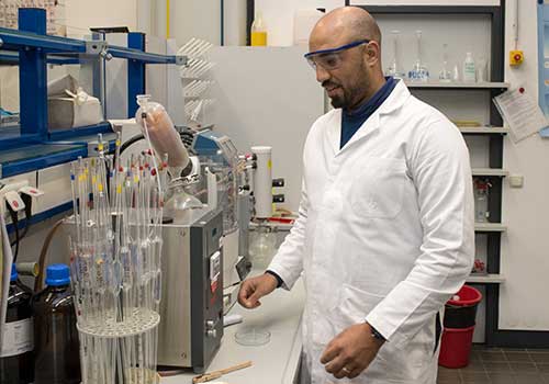 Ein lächelnder Mann in Kittel und mit Schutzbrille steht in einem Labor und führt ein Experiment durch.